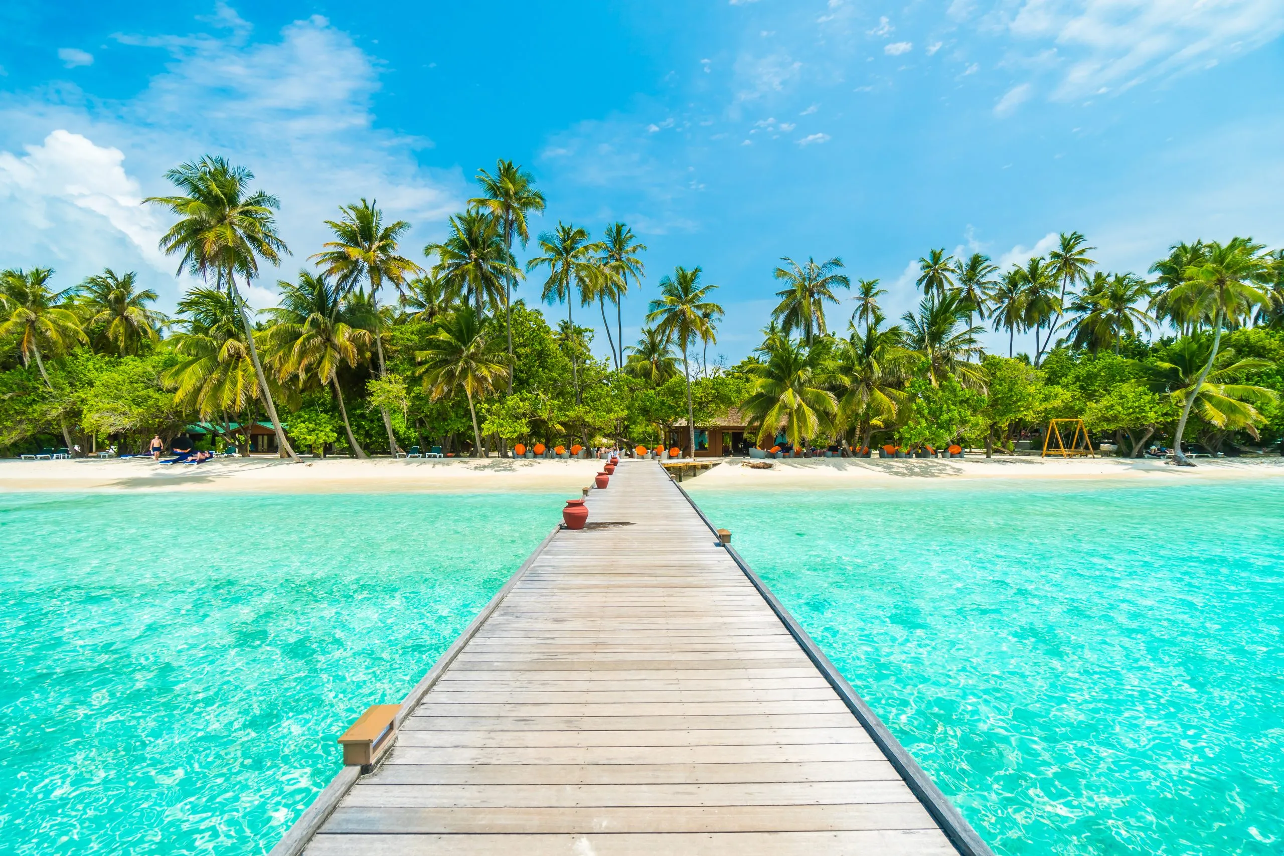 Un muelle de madera se extiende sobre aguas turquesas claras hacia una playa tropical bordeada de palmeras. El cielo es de un azul brillante con algunas nubes dispersas, lo que crea una atmósfera serena y acogedora.